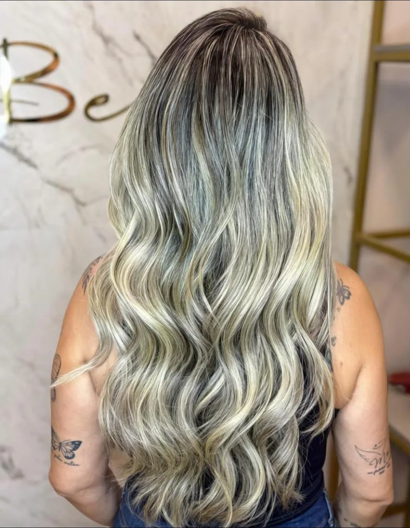 Cabelo longo com mechas loiras e ondas, visto de costas, diante da parede de mármore do salão